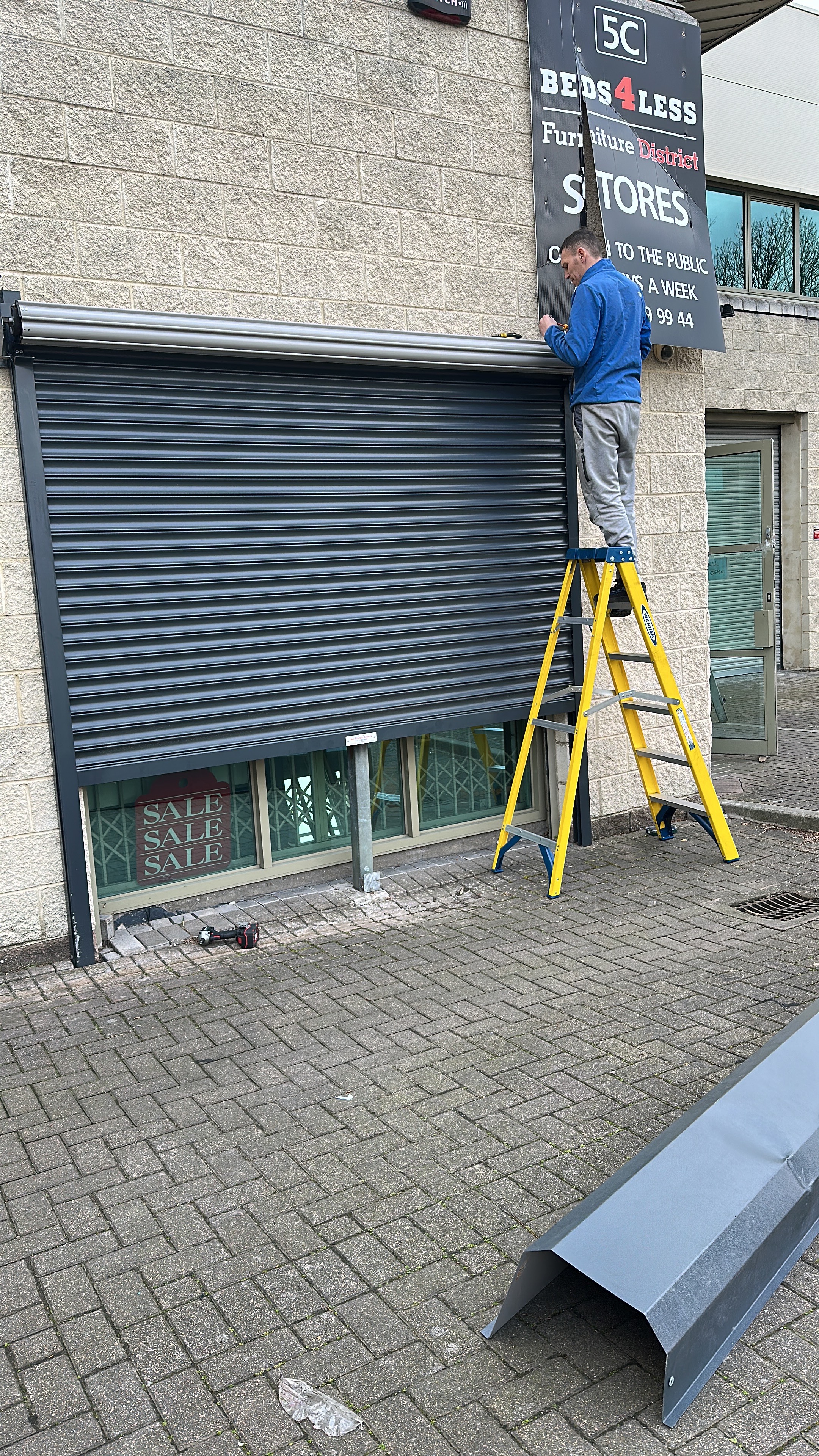 SteelMotion engineer on a ladder fitting a dark anthracite roller shutter to the Beds4Less furniture store shopfront in Dublin